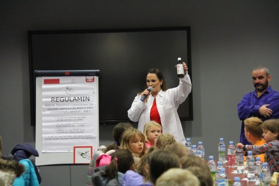 Obok stojaka z regulaminem warsztatów stoi prowadząca zajęcia pokazując dzieciom, butelkę oznaczoną substancjami niebezpiecznymi. Dzieci siedzą przy stołach