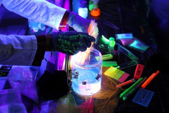 Do słoika z cieczą zanurzane są rurki. Zdjęcie zrobione po ciemku aby oddać fluorescencyjne właściwości przedmiotów