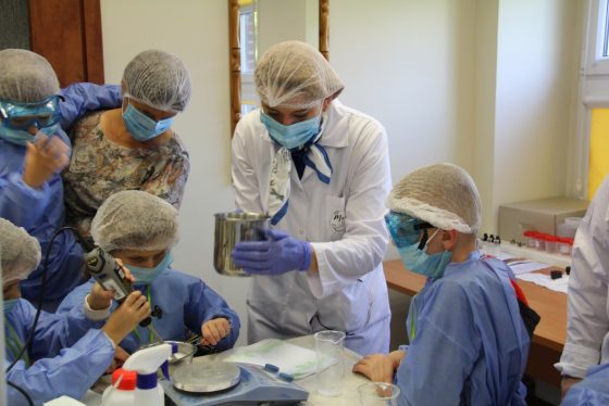 sześć osób w strojach ochronnych w laboratorium pochylonych nad doświadczeniem chemicznym. Jedna osoba w białym fartuchu donosi składnik, druga miesza składniki. Pozostałe osoby przyglądają się doświadczeniu